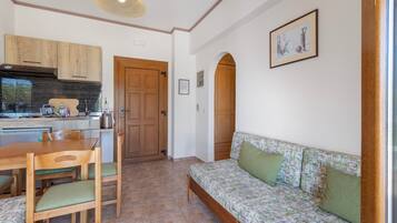 Ein gemütlicher Wohnbereich mit einer Küchenzeile, einem Holztisch mit Stühlen, einer Couch mit Kissen und einem gerahmten Bild an der Wand.