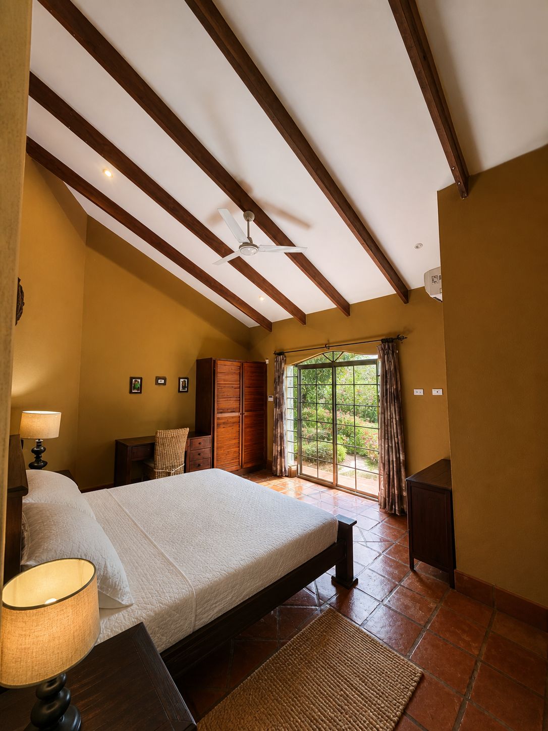 A bedroom with a bed, a nightstand, a lamp, a ceiling fan, and a view of greenery through a window.