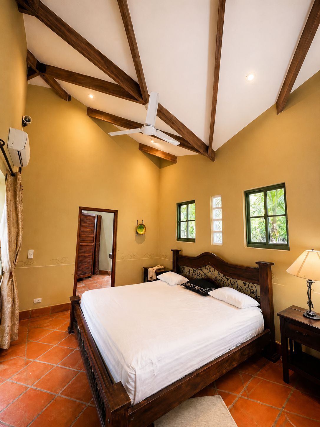 A bedroom with a wooden bed, a ceiling fan, a nightstand, a lamp, and a window with a view of greenery.