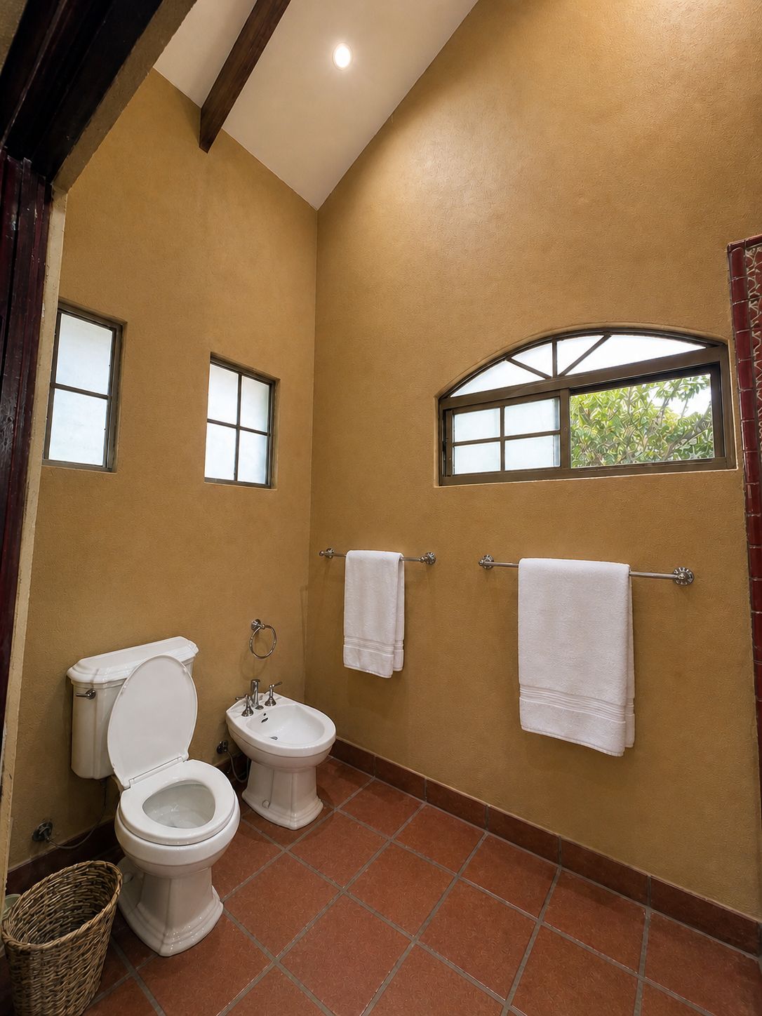 A bathroom with a toilet, bidet, and sink, two towels on towel racks, and a window with a view of trees.
