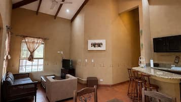 A spacious room with a high ceiling, wooden beams, a dining table with chairs, a sofa, a television, and a kitchen area with a bar.