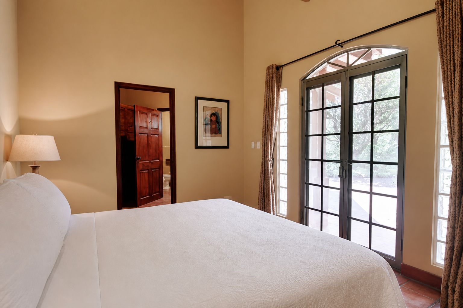 A bedroom with a large bed, a door leading to another room, a framed picture on the wall, and a window with curtains.