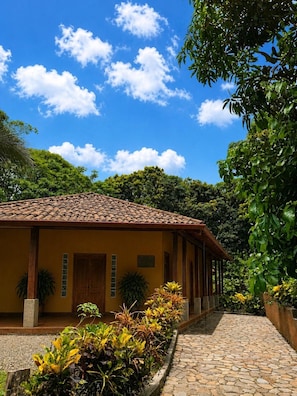 Un edificio con techo de tejas, acceso empedrado y un jardín con diversas plantas.