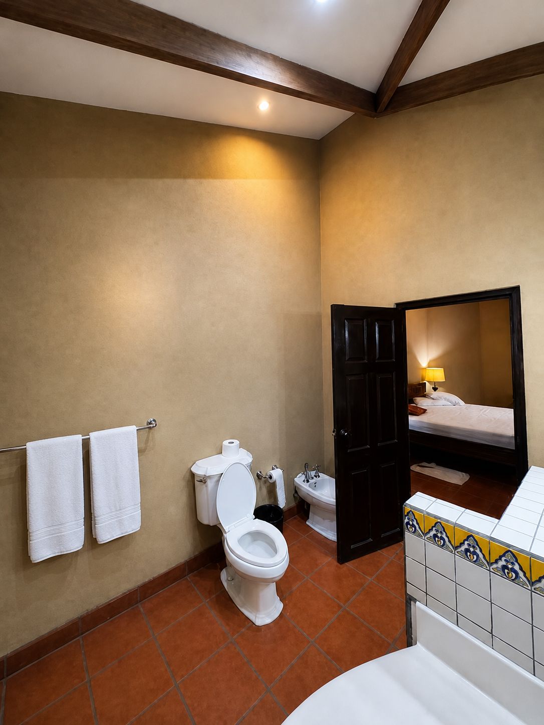 A bathroom with a toilet, bidet, and a sink. There are two towels on a rack, and a bed is visible through an open door.