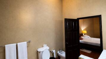 A bathroom with a toilet, bidet, and a sink. There are two towels on a rack, and a bed is visible through an open door.