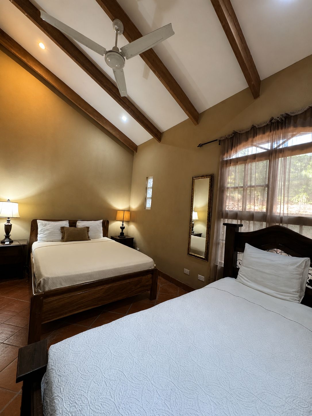 Une chambre avec deux lits, un ventilateur de plafond, une fenêtre avec des rideaux légers et un miroir au mur.