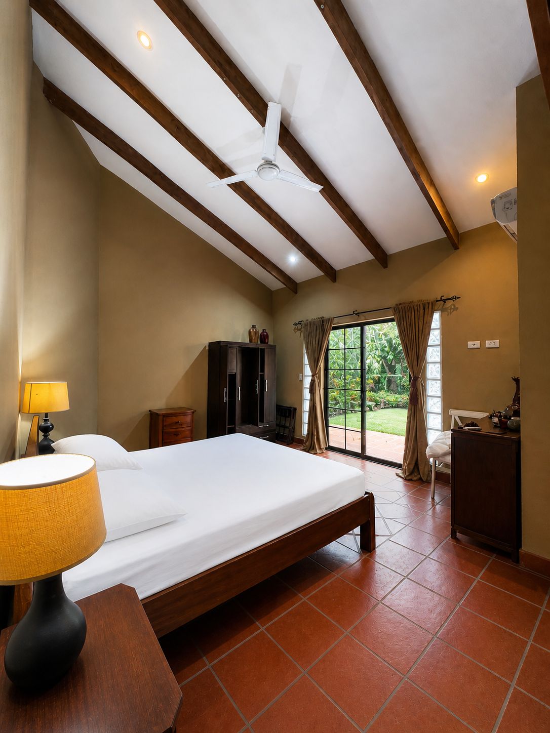 Un dormitorio con cama, mesita de noche, lámpara, ventilador de techo y vistas al exterior a través de una puerta de cristal.