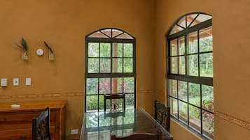 A dining area with a glass-top table, wooden chairs, a wooden cabinet, and two arched windows with views of greenery.
