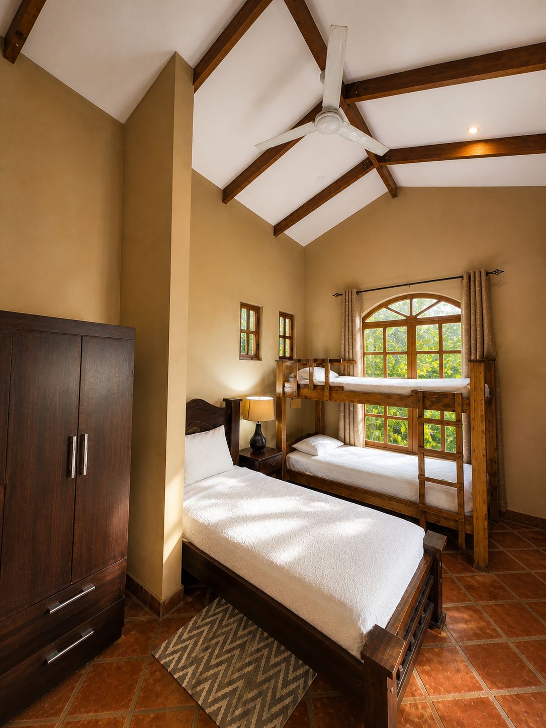 A bedroom with a large wardrobe, a single bed, two bunk beds, a ceiling fan, and a window with curtains.