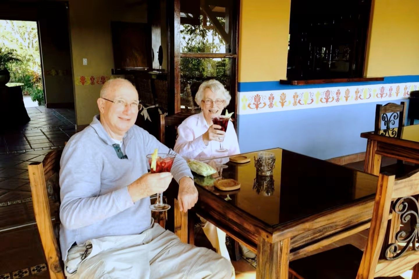 Two individuals seated at a wooden table with a glass of drink in each hand, in a room with a tiled floor and a window with a view of greenery outside.