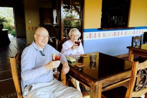 Two individuals seated at a wooden table with a glass of drink in each hand, in a room with a tiled floor and a window with a view of greenery outside.