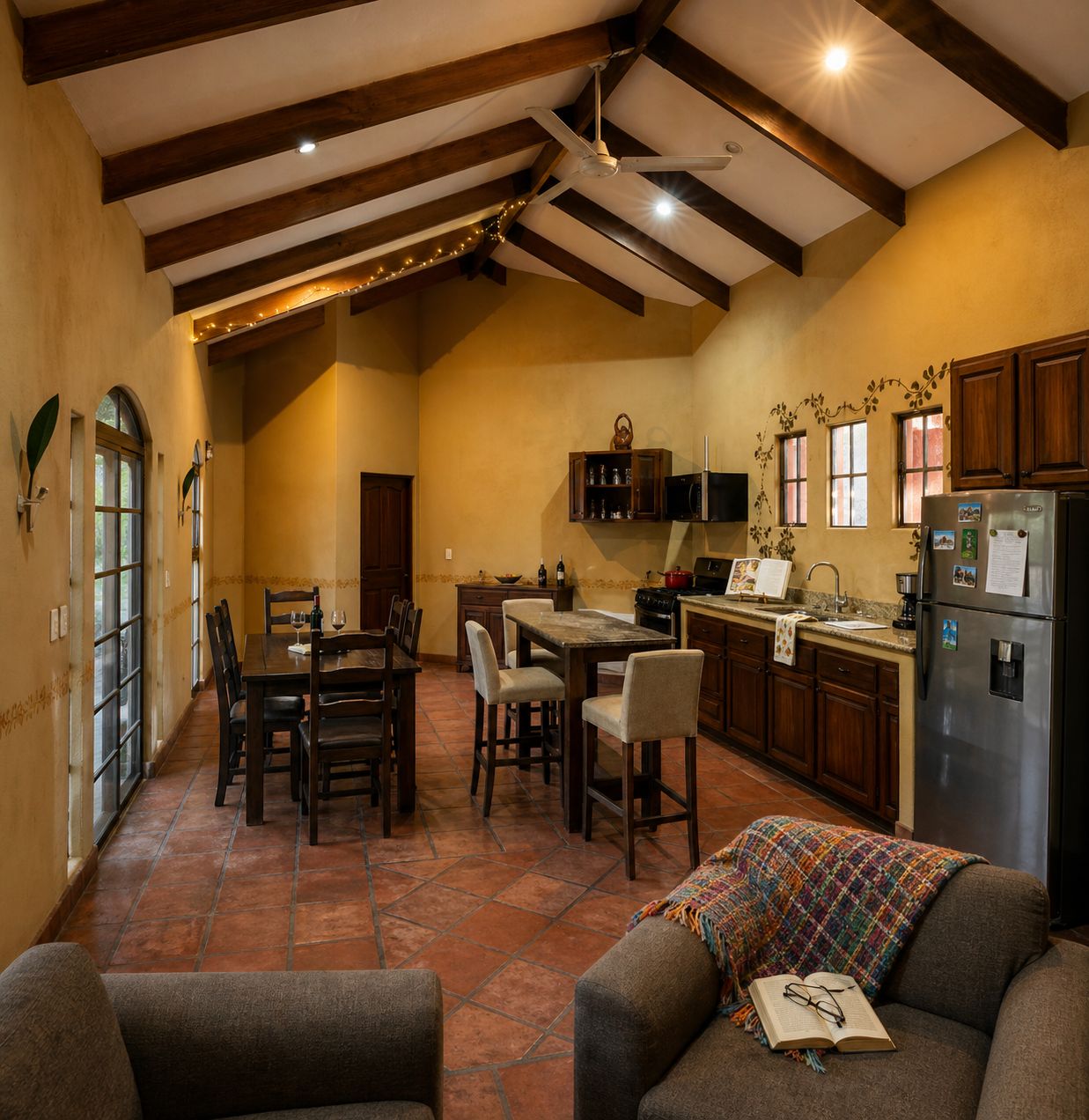 Una cocina amplia con vigas de madera, una zona de comedor con sillas, un sofá con una manta y un refrigerador.