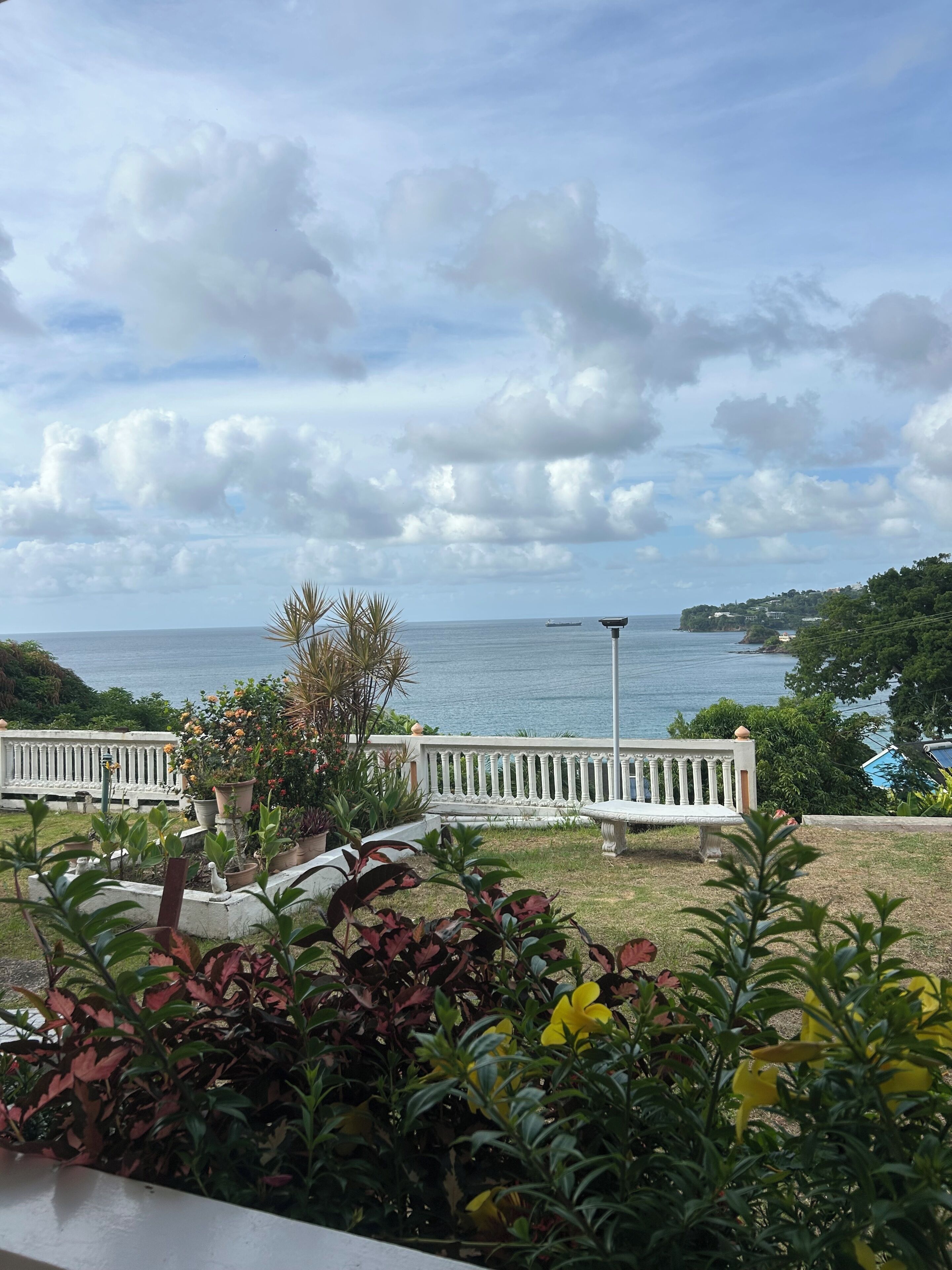 A view of a coastal area with a bench and potted plants in the foreground, a clear sky, and a distant view of the sea and a small island.