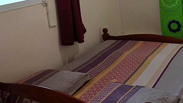 Une petite chambre avec un lit simple, une fenêtre avec des stores, un rideau rouge, un appareil de climatisation et un objet vert accroché au mur.