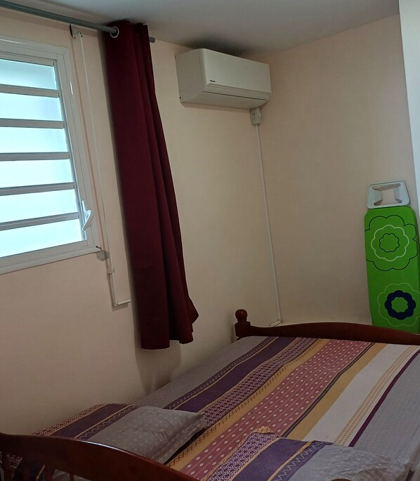 Een kleine slaapkamer met een enkel bed, een raam met zonwering, een rood gordijn, een airconditioning en een groen voorwerp aan de muur.