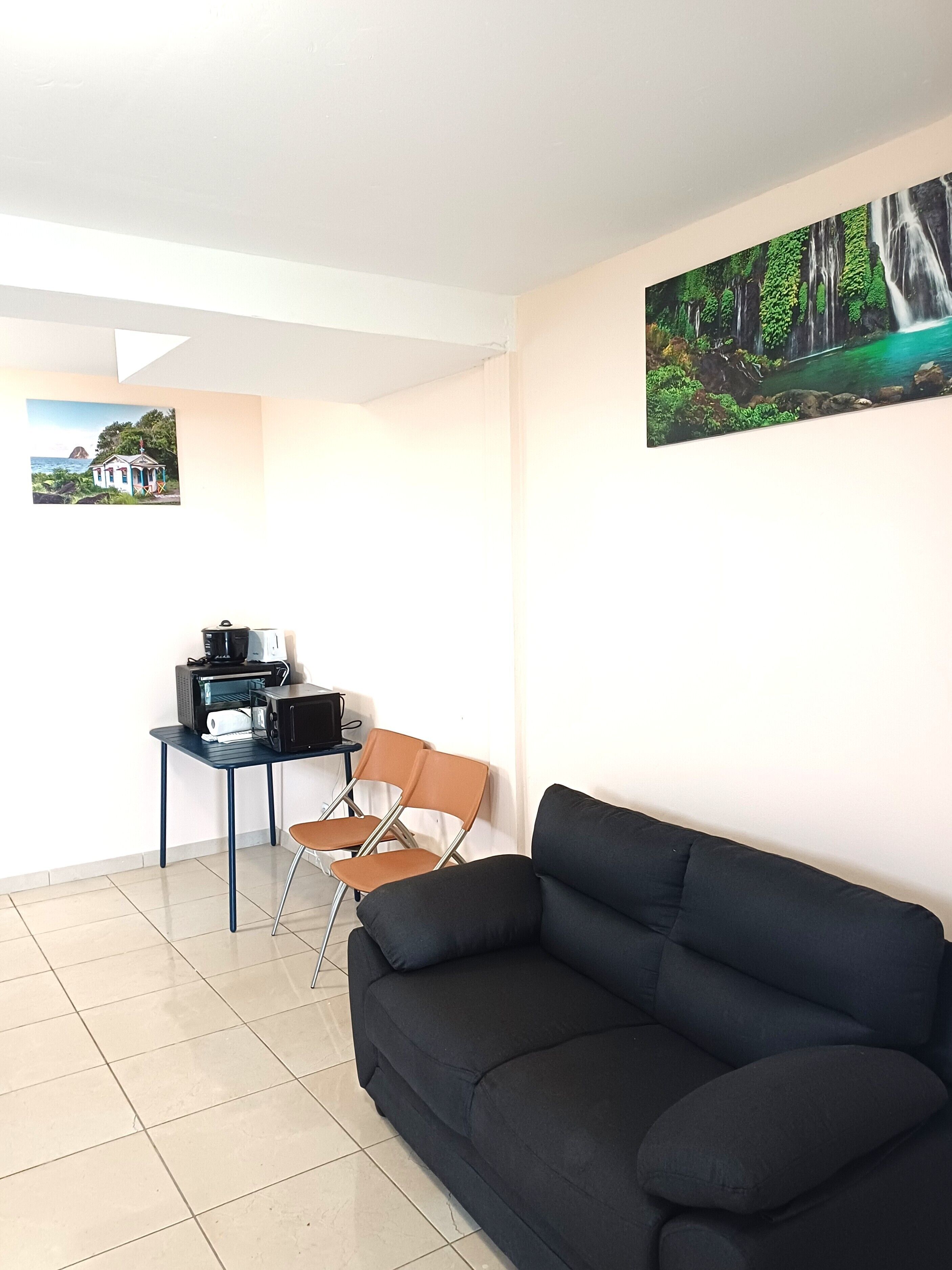 A room with a black sofa, a small table with a coffee maker, two chairs, and two framed pictures on the walls.