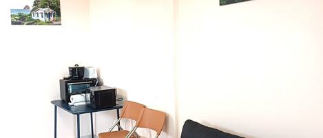 A room with a black sofa, a small table with a coffee maker, two chairs, and two framed pictures on the walls.