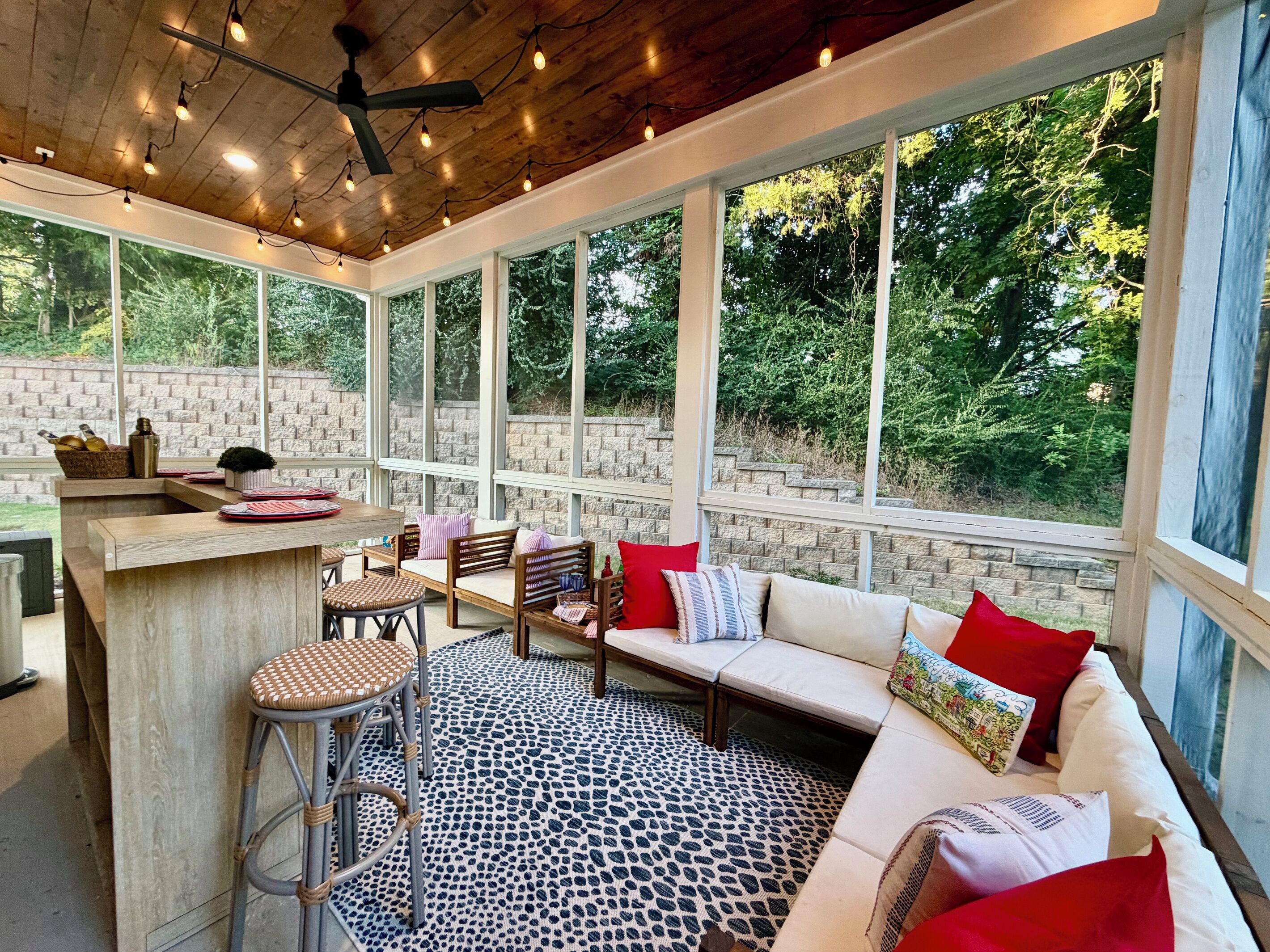 Un espace extérieur abrité doté d’un plafond en bois, de grandes fenêtres, d’un coin salon avec un canapé et des fauteuils, et d’un petit bar.