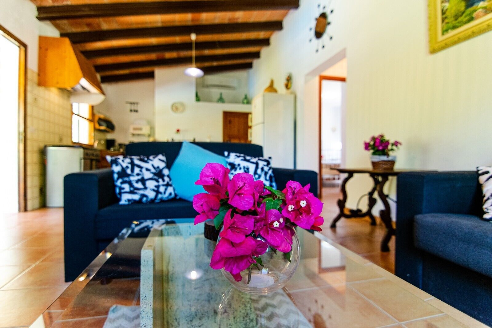 A living room with a glass coffee table, a blue sofa with patterned cushions, and a vase with pink flowers.