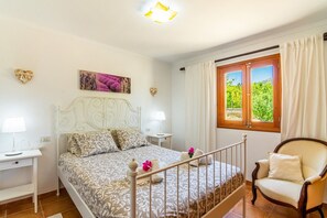 A bedroom with a bed, bedside tables, a chair, a window with curtains, and a painting on the wall. - Can Pep – Country House in Pollensa with Pool (Pollenca)