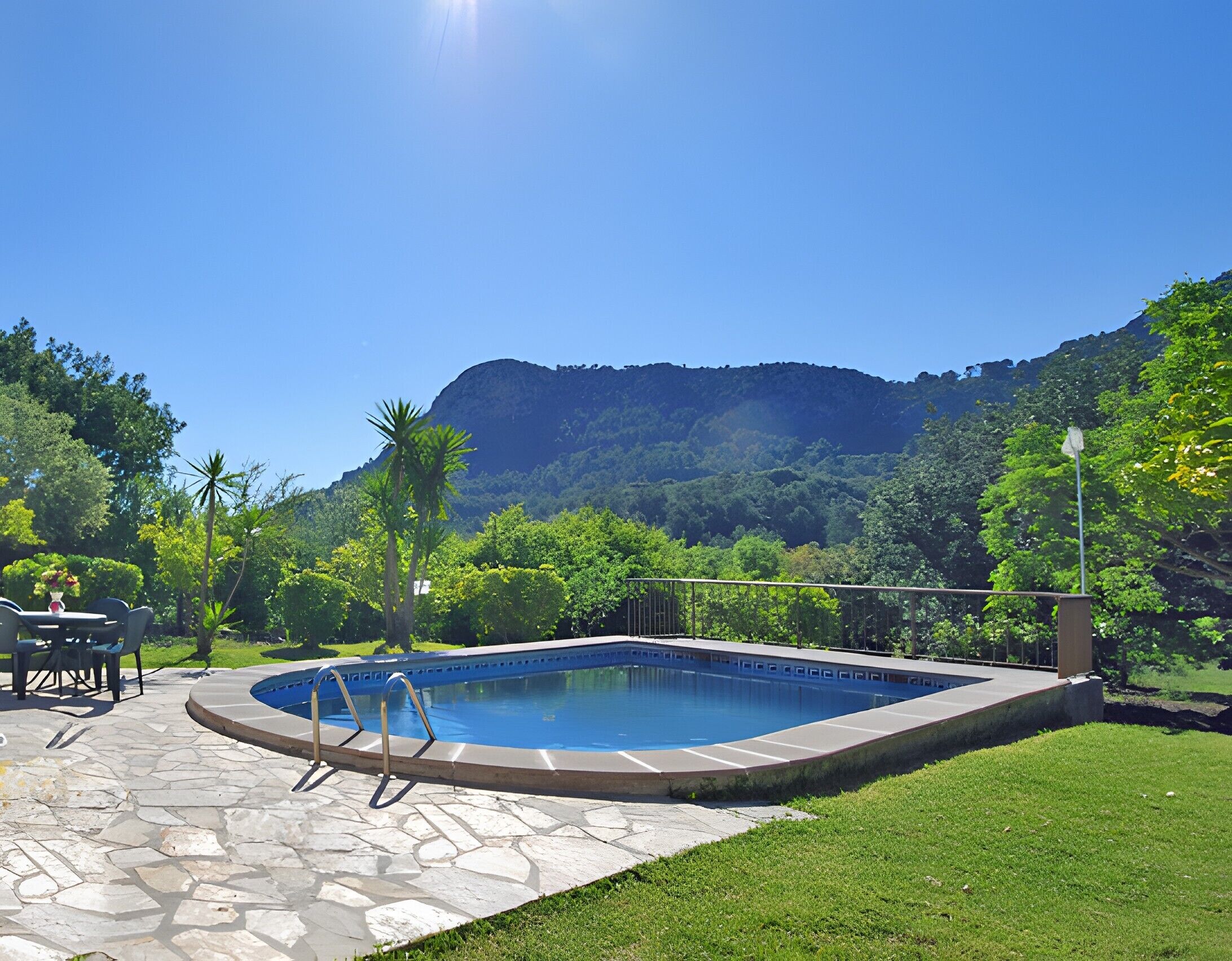 A swimming pool with a stone patio, surrounded by greenery and a mountain backdrop.