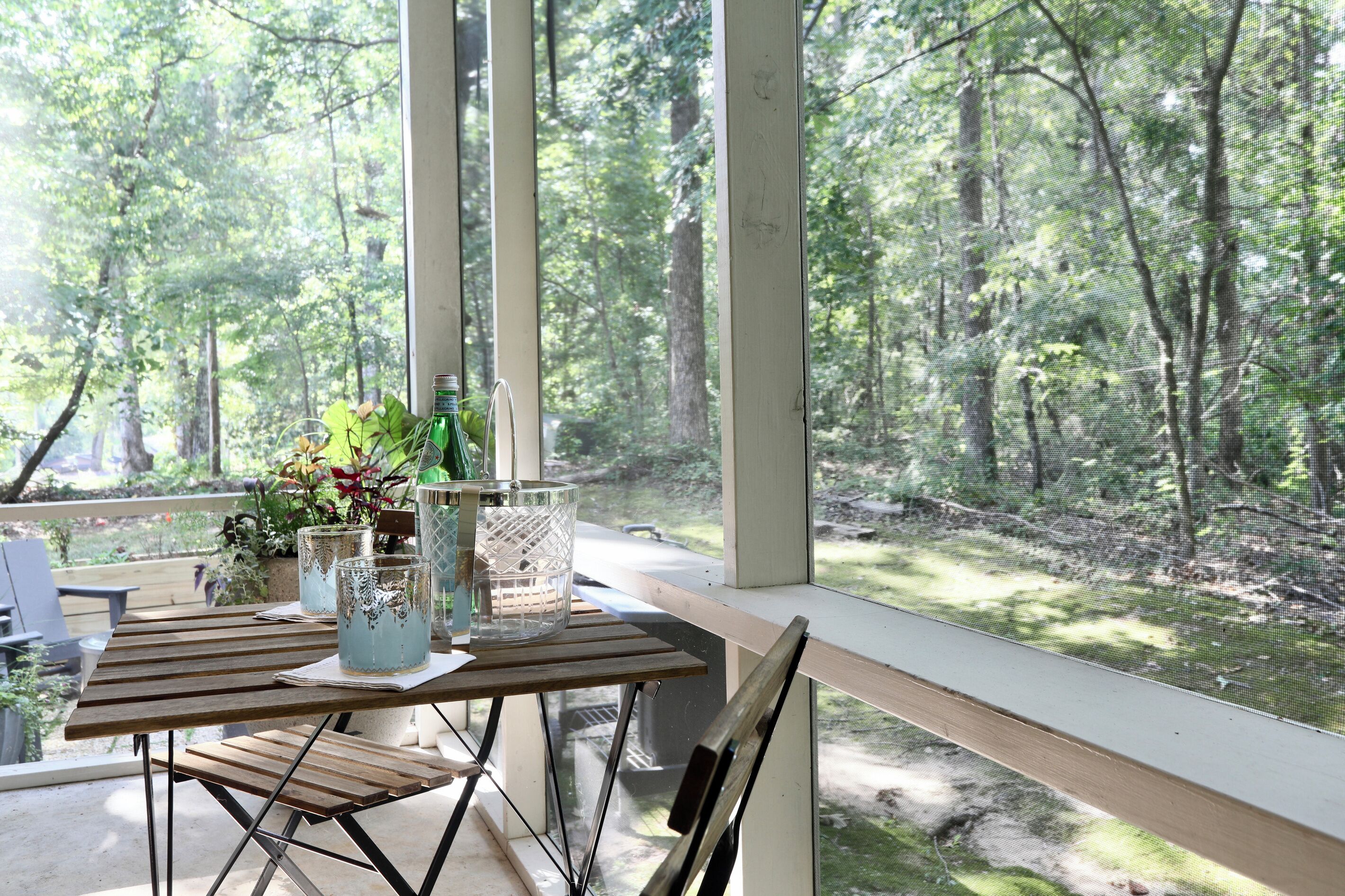 A wooden table with a bottle and glasses, a plant, and a decorative item on a balcony with a view of a forest.