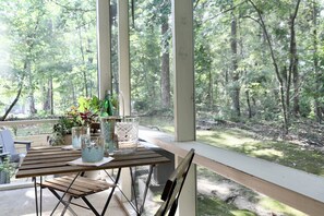 A wooden table with a bottle and glasses, a plant, and a decorative item on a balcony with a view of a forest. - "Hillside Hideaway" by OVR (Oxford)