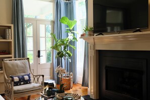 A living room with a fireplace, a television mounted above it, a bookshelf, a chair, and a large window with curtains. - "Hillside Hideaway" by OVR (Oxford)