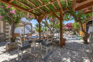 Eine Terrasse im Freien mit Steinboden, einer Holzpergola und einer Sitzecke mit Stühlen und Tischen.