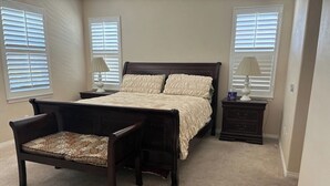 Un dormitorio con cama, una silla, una mesita de noche y dos ventanas con persianas.