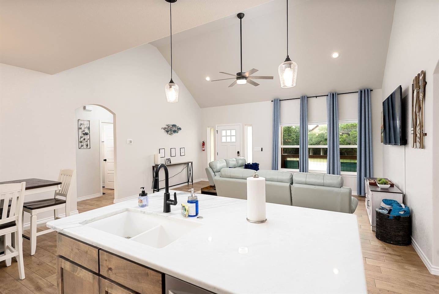 A modern kitchen with a large island, a dining area with white chairs, a grey sofa, and a ceiling fan.