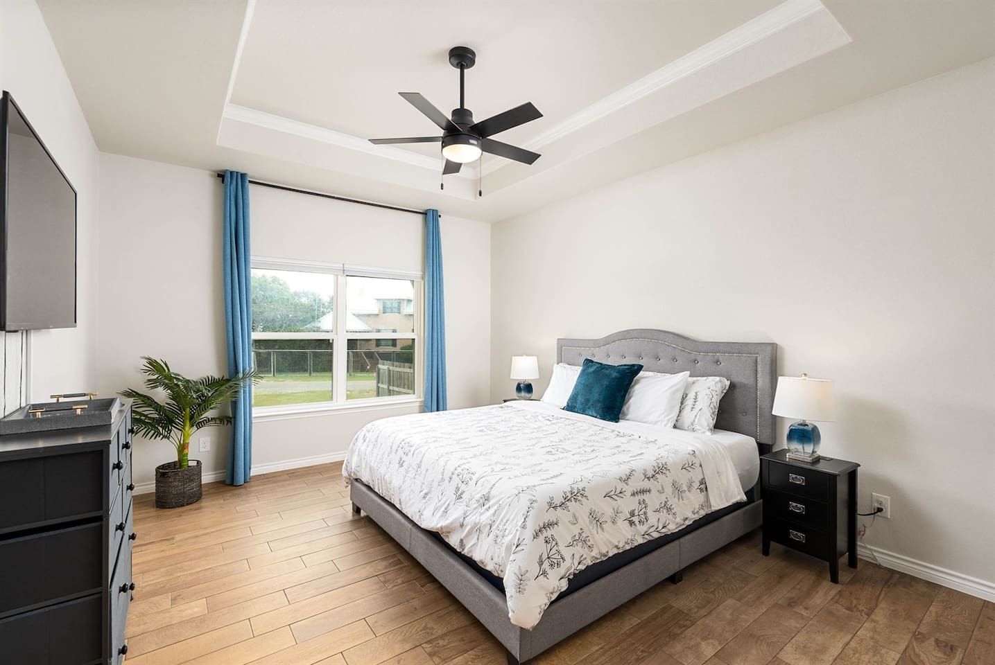A bedroom with a bed, a ceiling fan, a window with curtains, a nightstand with a lamp, and a plant.