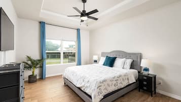 A bedroom with a bed, a ceiling fan, a window with curtains, a nightstand with a lamp, and a plant.