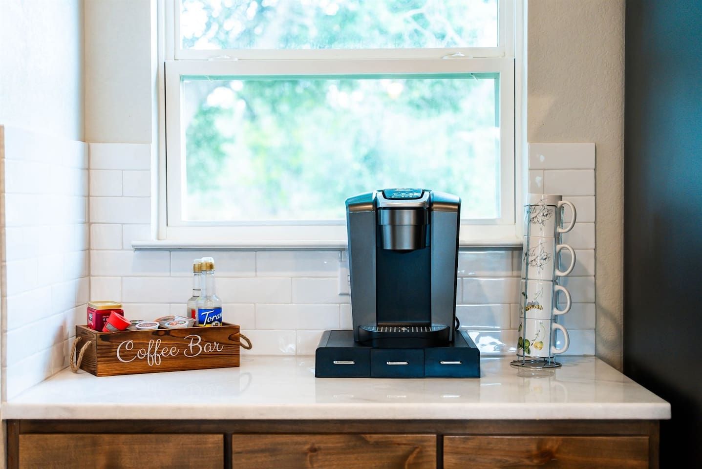 A coffee maker on a kitchen counter with a window above it, a coffee bar sign, and a mug rack.
