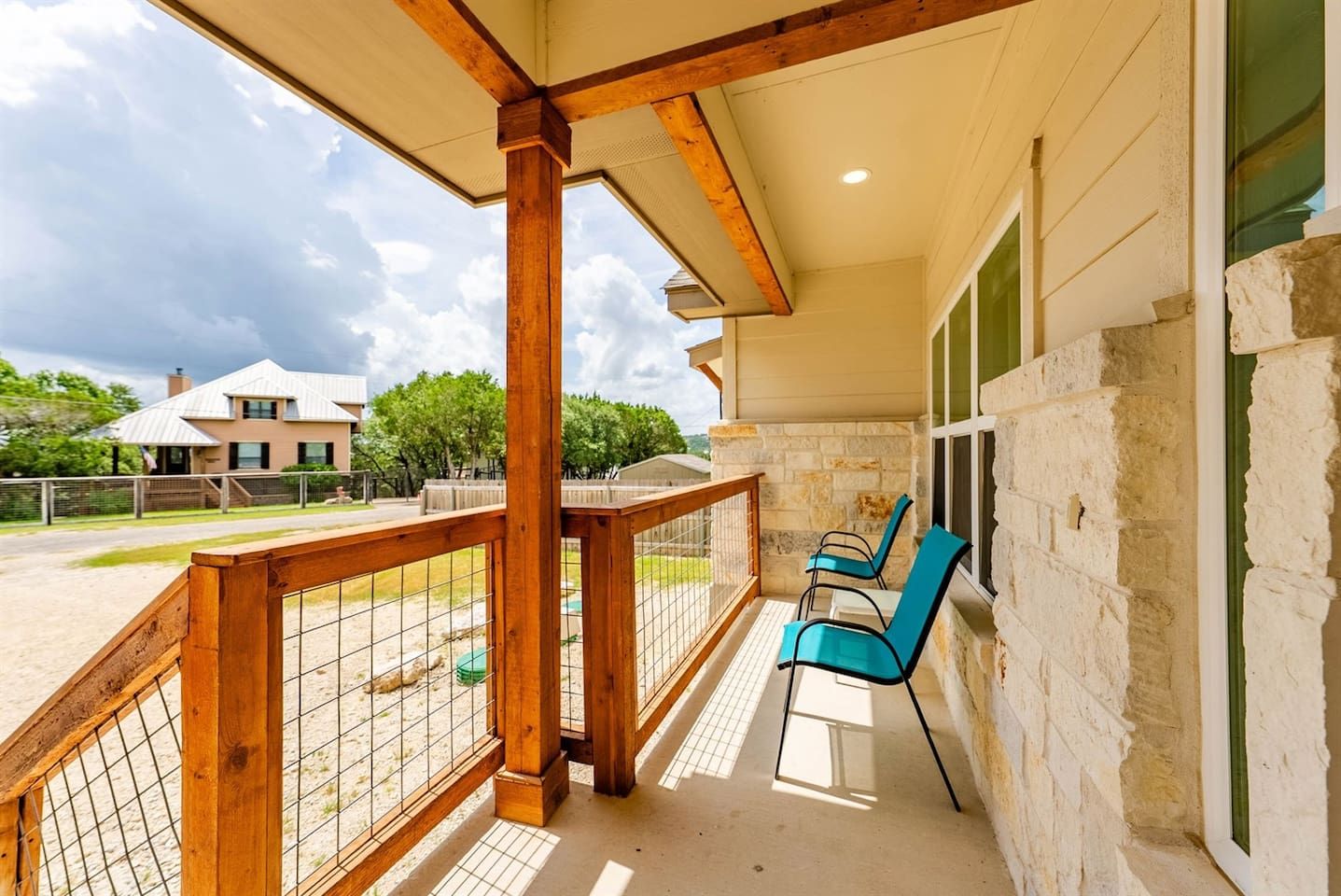 Un espacio al aire libre techado con barandal de madera, dos sillas y vista a una casa al fondo.