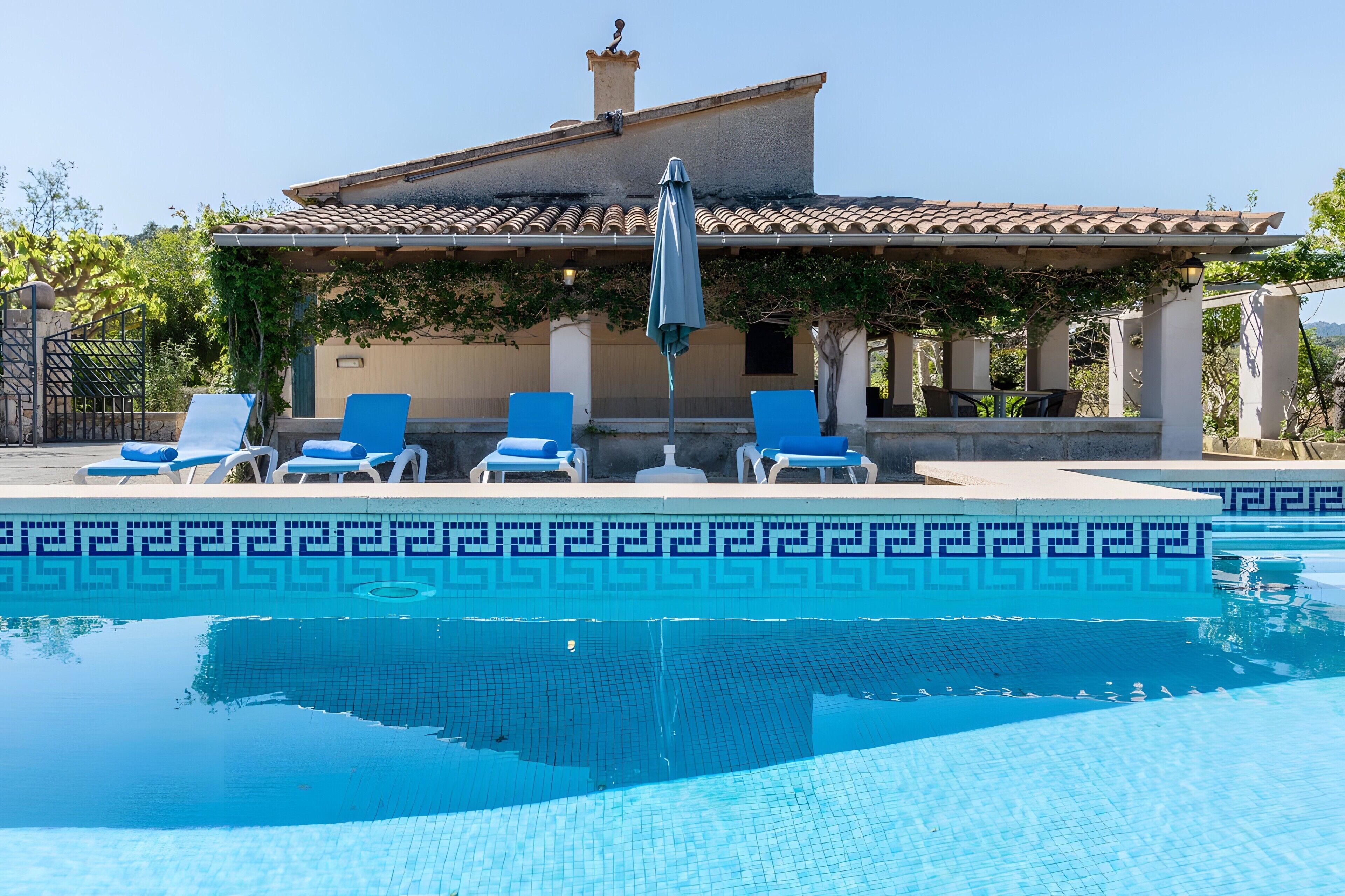 A pool with a geometric tile pattern, surrounded by lounge chairs and a building with a tiled roof.
