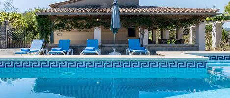 A pool with a geometric tile pattern, surrounded by lounge chairs and a building with a tiled roof.