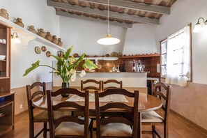 A dining area with a round wooden table and chairs, a hanging light fixture, and shelves with pottery and plates. - Holiday Home "Catalina Petit" with Mountain View, Pool, Garden, Terrace & Wi-Fi (Pollença)