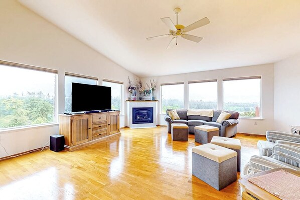 A spacious living room with a flat-screen TV, wooden flooring, a fireplace, a gray sofa, and large windows offering a view of the outdoors.