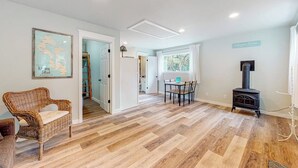 Una stanza con pavimento in parquet a spina di pesce, una poltrona in vimini, un tavolo da pranzo con sedie, una stufa a legna in legno nero e una finestra vista alberi.