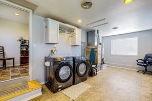 A laundry room with two front-loading washers and dryers, a cabinet above the dryers, a window with blinds, and a chair. - Charming Single Level Home with Fireplace, Patio, and Fast WiFi in Oak Harbor (Oak Harbor)