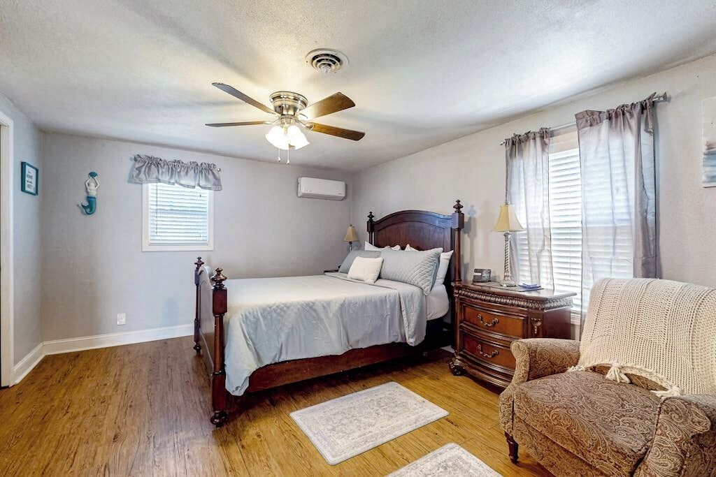 Quarto com cama, cômoda, cadeira, ventilador de teto e janela com cortinas.
