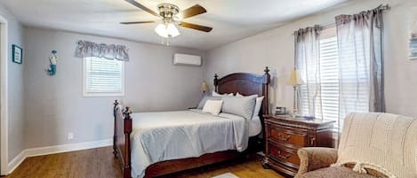 Une chambre à coucher comprenant un lit, une commode, une chaise, un ventilateur de plafond et une fenêtre avec des rideaux.