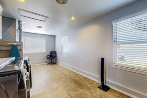A room with a kitchenette, a chair, a standing fan, and a window with blinds.