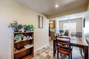 A dining area with a wooden table and chairs, a bookshelf with plants, and a kitchen in the background. - Charming Single Level Home with Fireplace, Patio, and Fast WiFi in Oak Harbor (Oak Harbor)