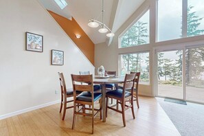 Un comedor con mesa y sillas de madera, una obra de arte en la pared y grandes ventanales con vistas a árboles.