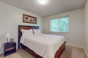 Un dormitorio con cama, mesita de noche, lámpara, cuadro enmarcado y ventana con vistas a árboles.