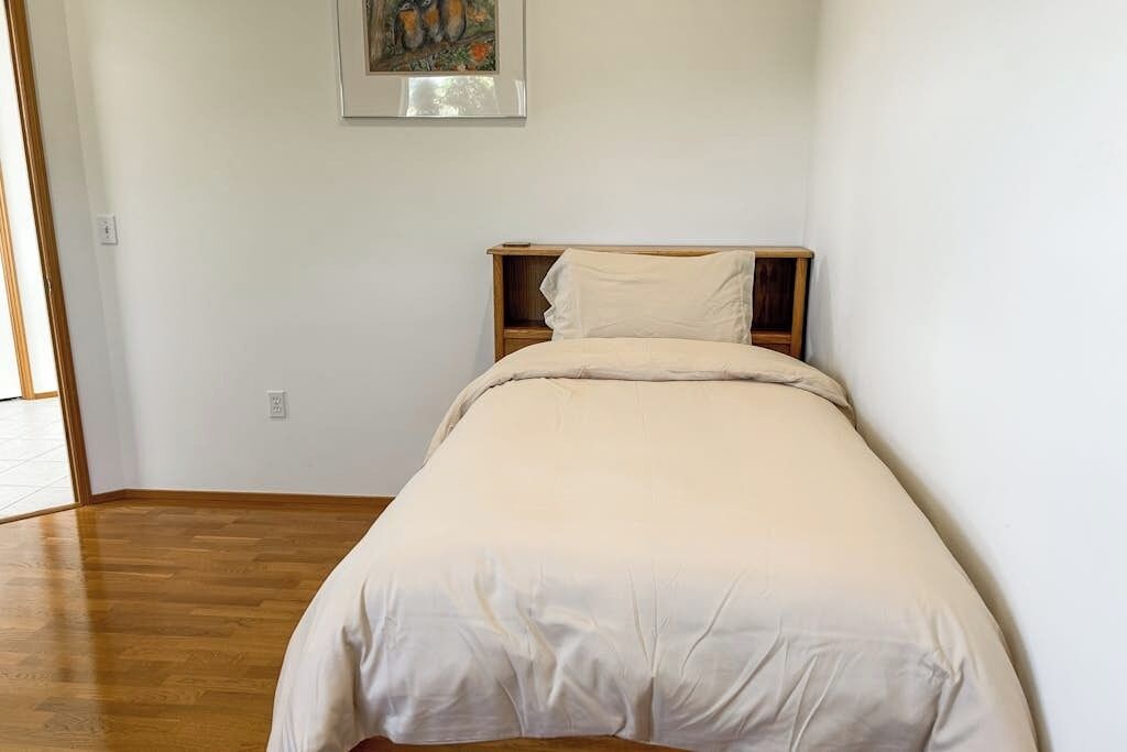 Un lit simple avec une tête de lit en bois, dans une pièce aux murs blancs et au plancher en bois.