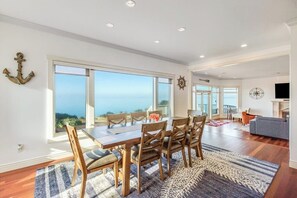 A spacious dining area with a large table and chairs, a view of the ocean through large windows, and a cozy living area with a fireplace. - Stunning Dog Friendly Waterfront Villa with Sunset Views and Fireplace (Oak Harbor)