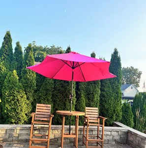 Une terrasse avec deux chaises en bois et une table, un parasol rouge et un mur de pierre agrémenté de verdure en arrière-plan.